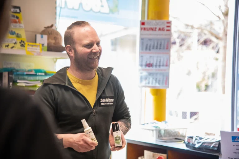 Mann mit Bart hält zwei Flaschen in einem Geschäft, lächelnd und in einem grünen Sweatshirt.